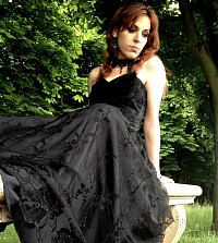 Portrait d’Elsy Eala, autrice de romans fantastiques, vêtue d’une robe noire élégante, assise sur un banc de pierre dans un parc verdoyant.